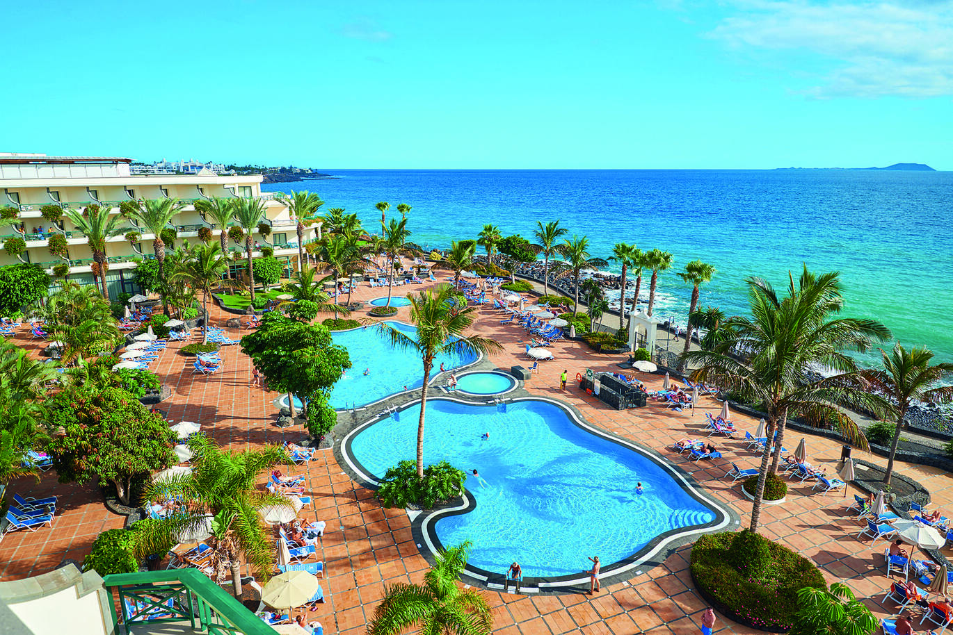 Aerial view of the pool area surrounded by sun loungers, palm trees, people in and around the pool with part of the hotel building visible behind. The sea is directly behind with the blue, slightly cloudy sky above.