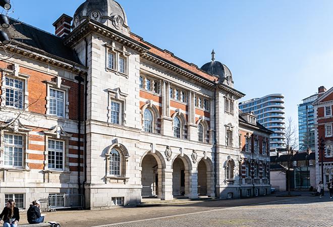 “Historic building with red brick and white stone facade featuring arched entrances and decorative domes, located in an open courtyard with modern high-rise buildings visible in the background under a clear blue sky.”