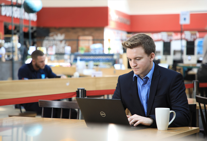"Photograph of a white male sitting at a table with his laptop with a cup beside him"