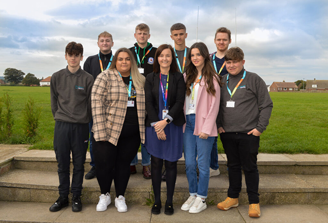 "Photo of 9 people, who are Beyond Housing apprentices, standing on 2 steps with a field in the background, smiling for a photo. Each wearing a Beyond Housing lanyard."