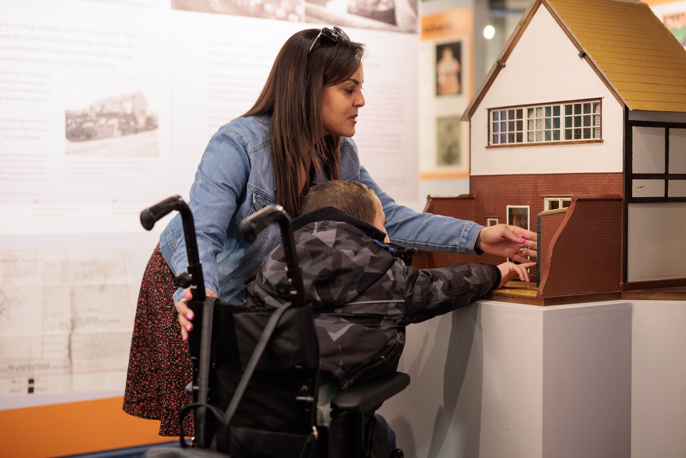 A woman assists a child in a wheelchair as they interact with a detailed model building at a museum exhibit.