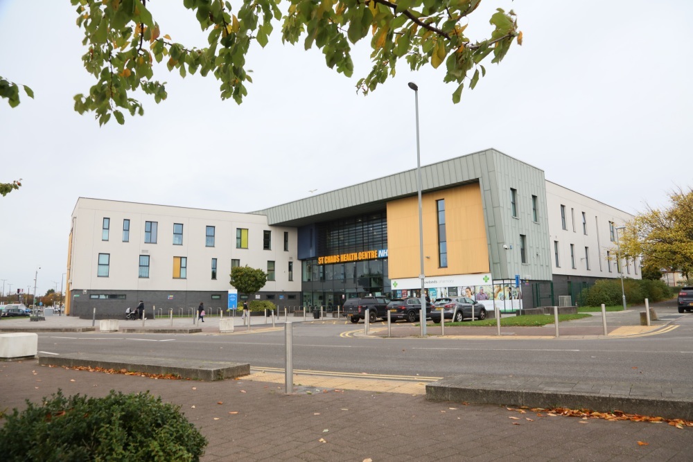 "A large 3 storey building with its name in large orange letters which reads ' St Chads Health Centre NHS'. There is a small car park outside the front of the building and a pharmacy on the ground level."