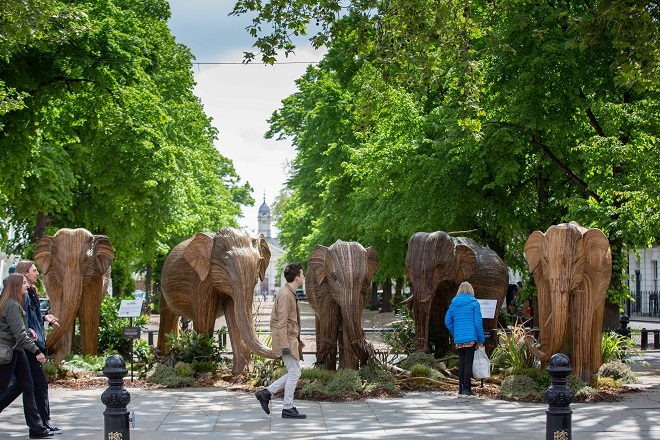 "Photo of 5 art display 3D elephants with people walking on the pavement near them"