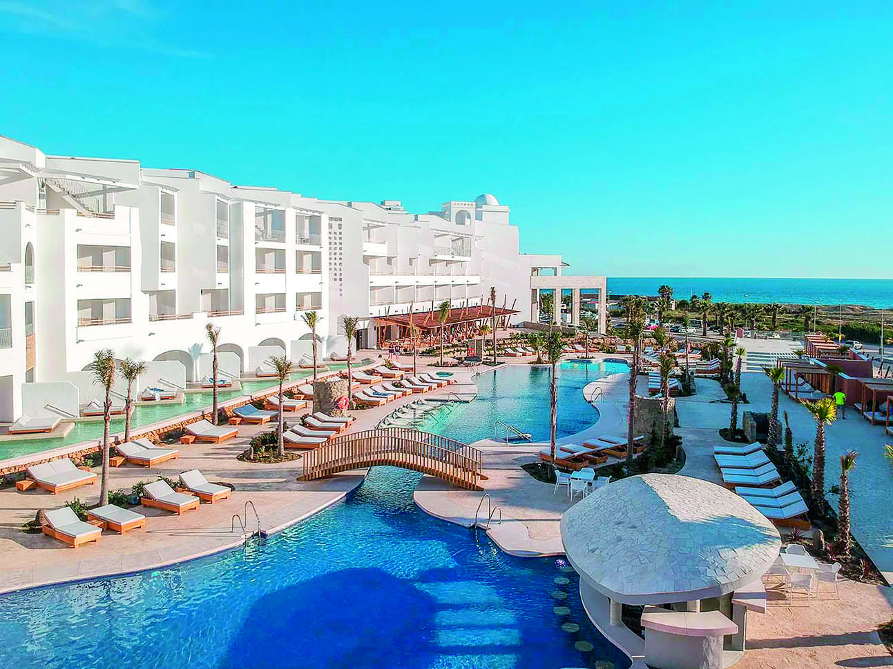 View of the pool surrounded by sun loungers with a little bridge over the pool with the hotel building to the left and the sea in the background on a clear, sunny day.