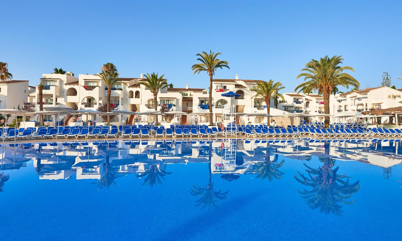 A view of the large, blue swimming pool surrounded by many blue sun loungers and palm trees behind. Behind this is the hotel building where you can see a cluster of hotel rooms with balconies. This is all also reflected in the pool. The sky above is clear and blue.