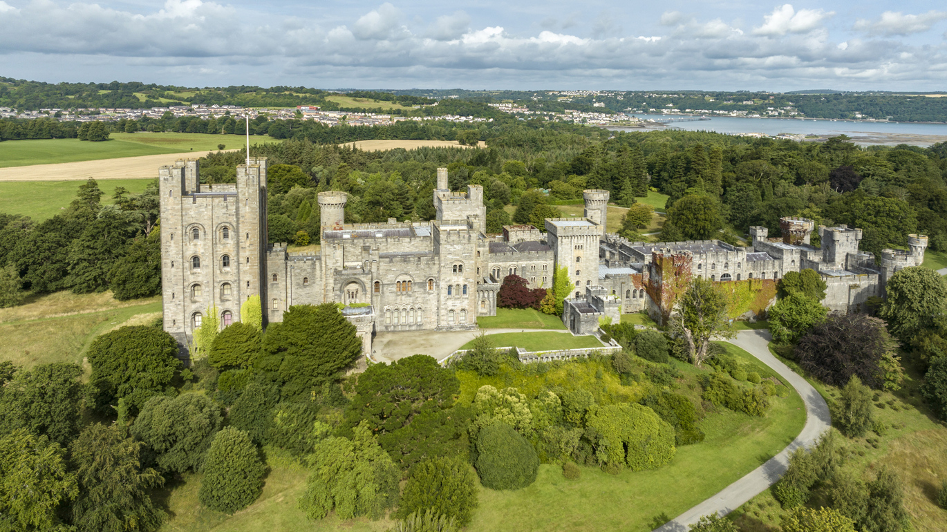 Aerial view of the 19th century Penrhyn Castle and Garden. Surrounded by lots of lawns and trees with buildings and a lake far in the background.