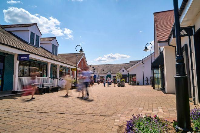 "Open-air outlet shopping village with white storefronts, brick paving, and blurred shoppers walking along a sunny street"