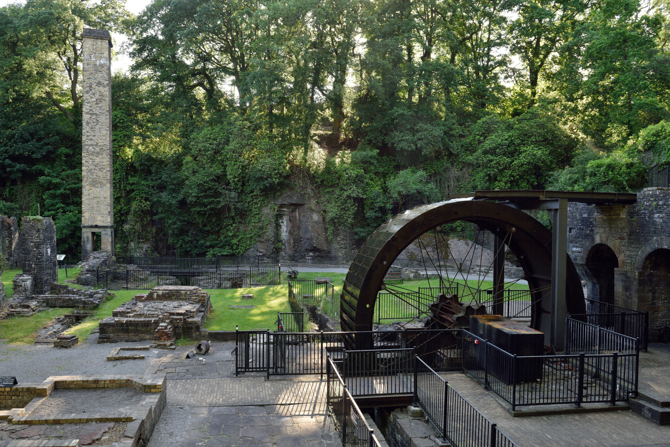 The waterwheel and bastion at Aberdulais, surrounded by brick ruins with a wall around it all with many trees and greenery up and around the wall.