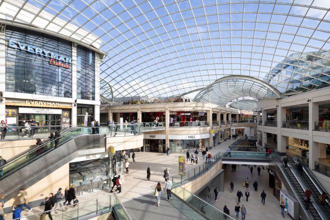 "Modern shopping centre interior with glass roof, multiple levels, escalators, and people walking between shops."