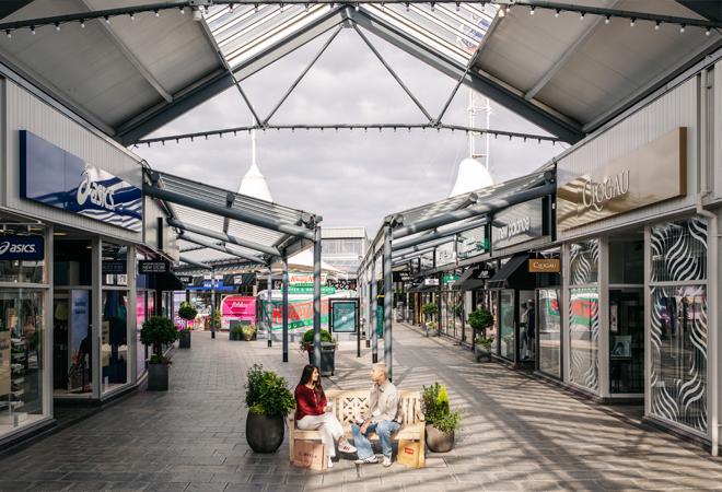 "Covered outdoor shopping walkway with retail stores and two people seated on a bench surrounded by potted plants."