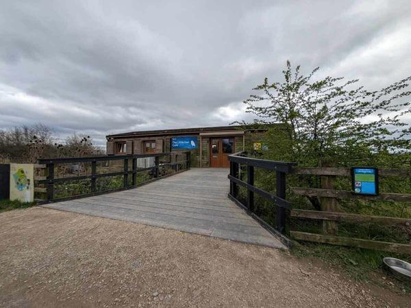 A wooden walkway with railings leads to a low visitor centre building with signage, surrounded by vegetation under a cloudy sky