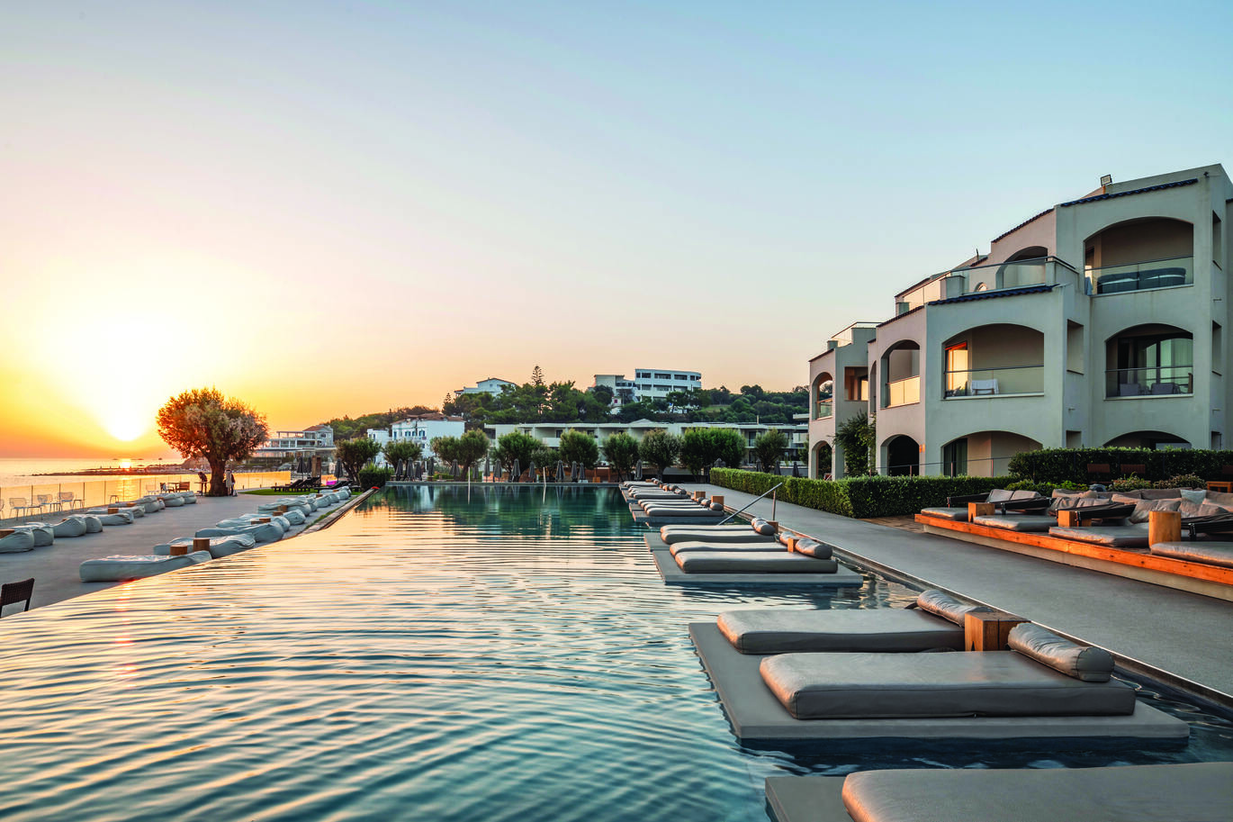 View of the pool area in the sunset with sun loungers in and around the pool and the hotel buildings behind.