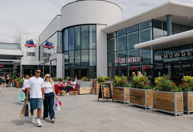 "Modern outlet center with glass-fronted shops, outdoor seating, planters, and people walking with shopping bags"