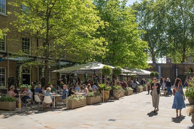 Photo of people sitting at tables and chairs outside restaurants under canopies and people walking around the path outside of the seating area