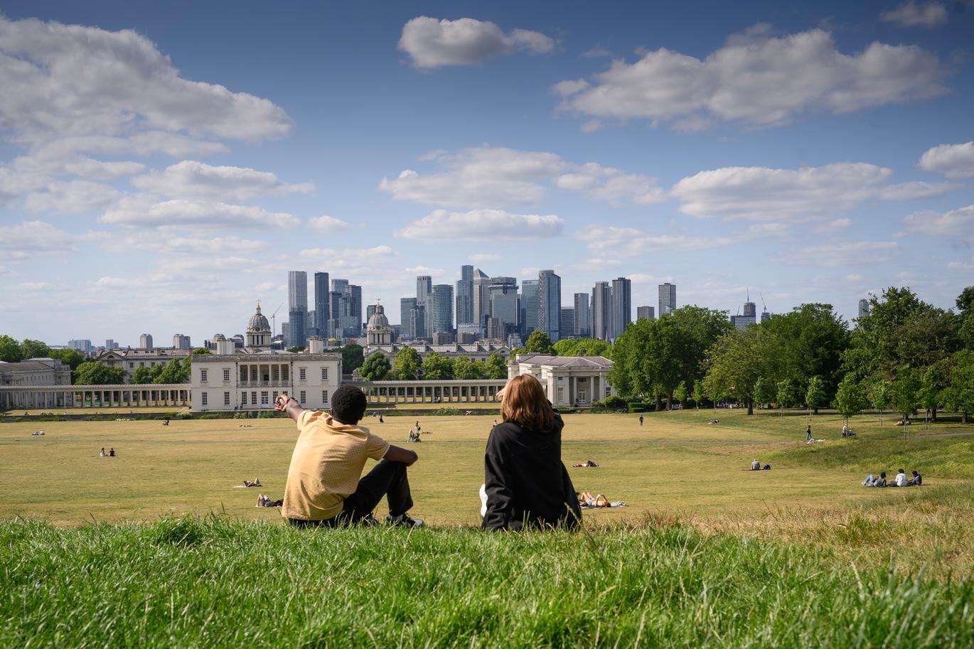 "Two people sit on a grassy hill in a large park, looking out towards a row of classical buildings with colonnades and a modern city skyline beyond. Other people are scattered across the lawn on a bright day under a partly cloudy sky"