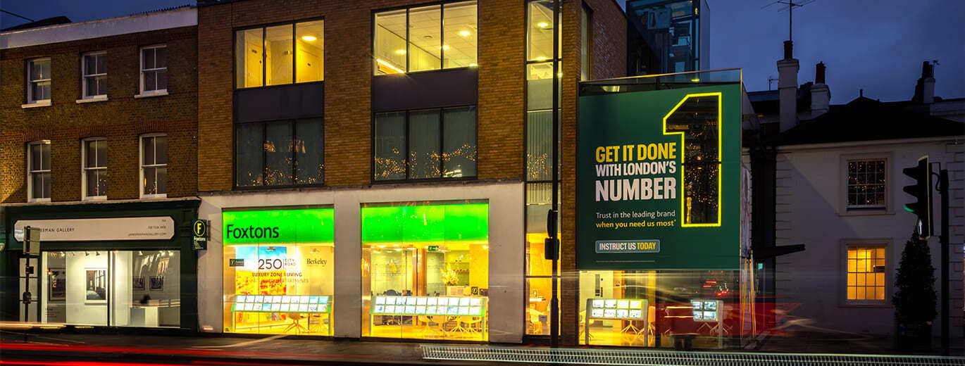 "The image captures a contemporary urban scene showcasing a modern building featuring a real estate agency, enhanced by illuminated storefronts and a prominent billboard advertising a service in London."