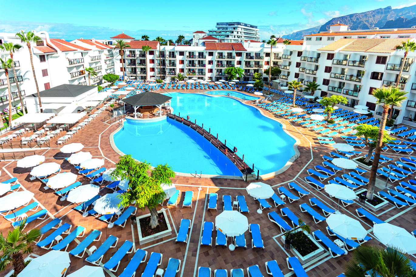 Aerial view of the pool area with a bridge over the middle surrounded by many blue sun beds and white umbrellas with the hotel building surrounding and mountains just visible in the background.