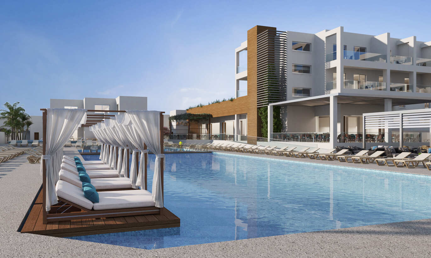 View of the pool area with sun beds undercover on the pool edge and more sun beds surrounding. The hotel building is visible behind with the blue sky above.