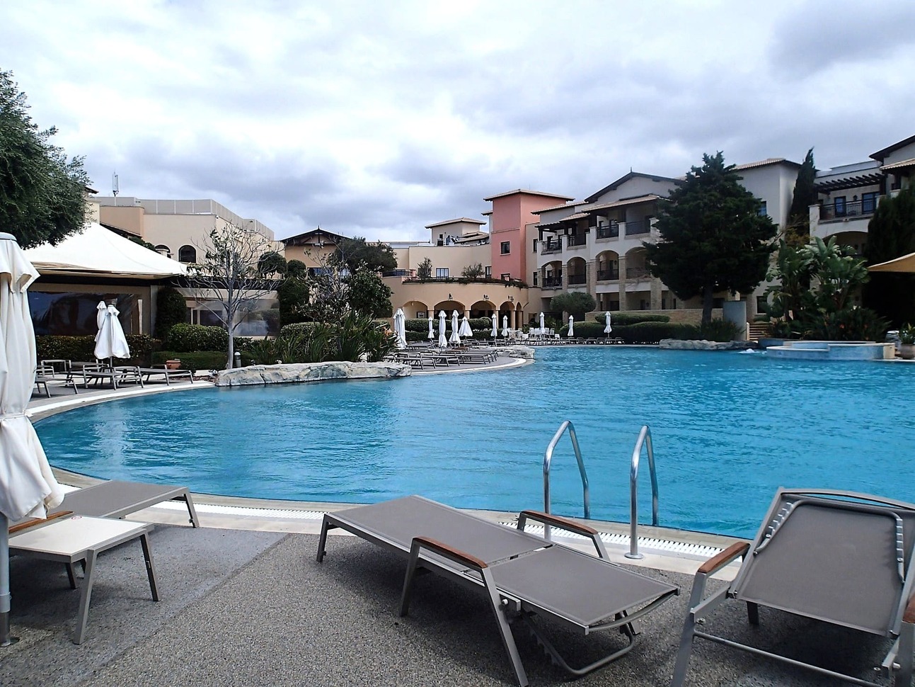 TUI BLUE Sensatori Atlantica Aphrodite Hills blue pool with hotel rooms in the background, some green trees around the pool and grey sun beds with white umbrellas around the pool.