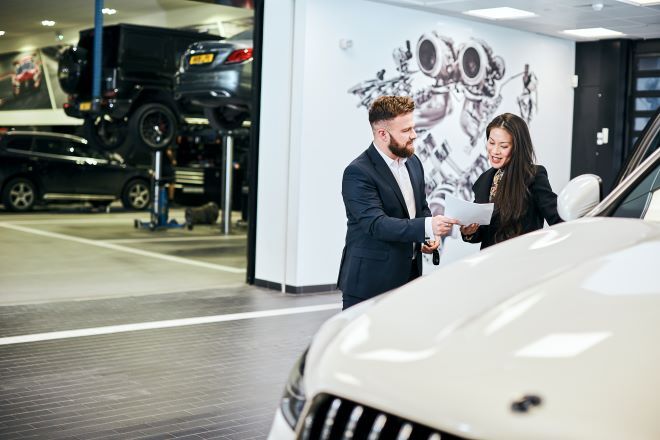 "two people are looking at paperwork behind a white car. in the background is a workshop with raised cars"