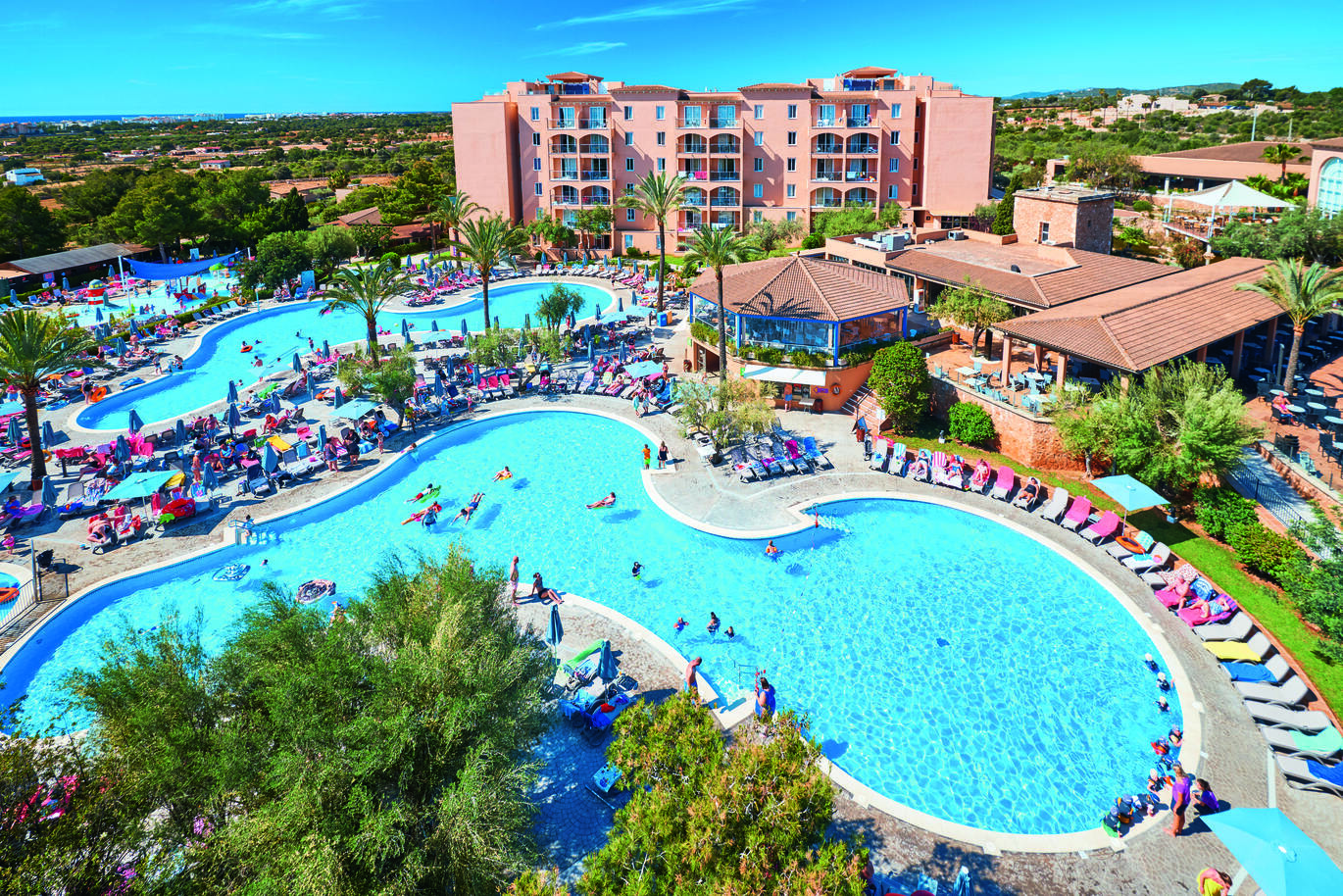 An aerial view of the 2 pools, one larger and one smaller, both surrounded by many sun loungers, umbrellas and people. You can see the hotel building behind and a smaller building in front. It is surrounded by plenty of greenery.