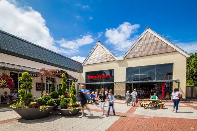 "Clarks Village walkway with shoppers outside stores including Superdry, with planters and seating areas."