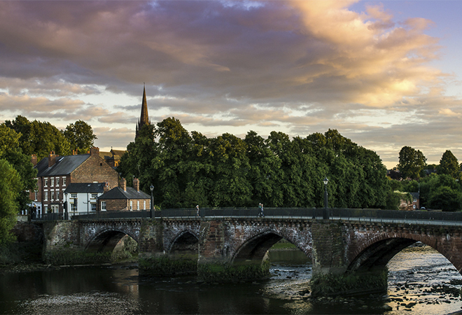 "Bridge over a river where a spire can be seen in the background with the sun setting behind the trees"