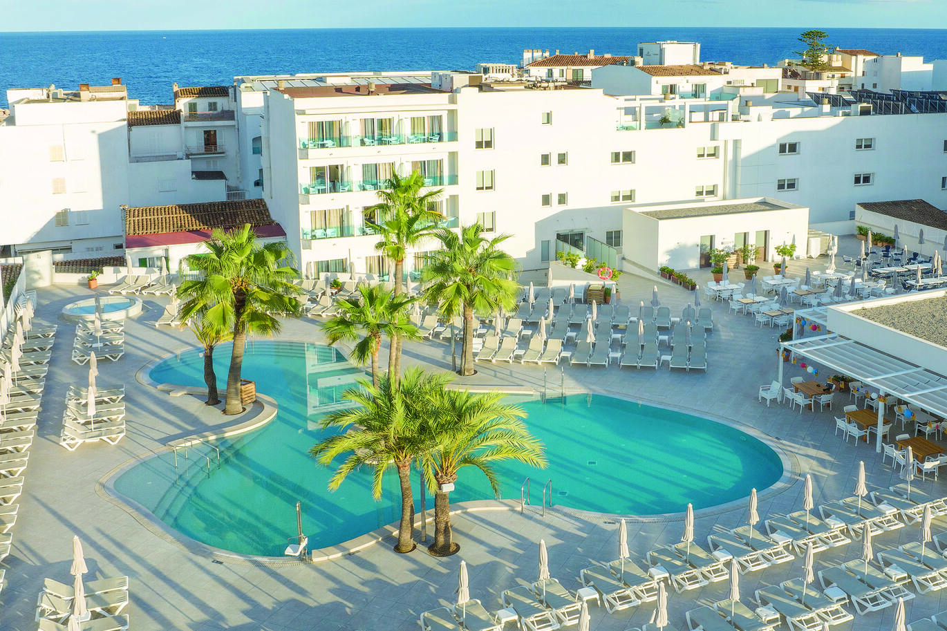 Aerial view of the pool area surrounded by sun loungers and palm trees dotted around the edge of the pool. With the hotel building behind and the sea just visible behind the building.