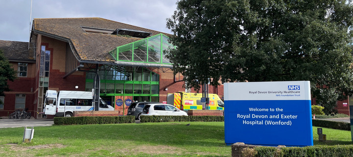 Blue 'welcome' signage in the foregound on the right, the multistorey hospital in the background. Large green windows on the front of the building and ambulanced parked outside the entrance. 