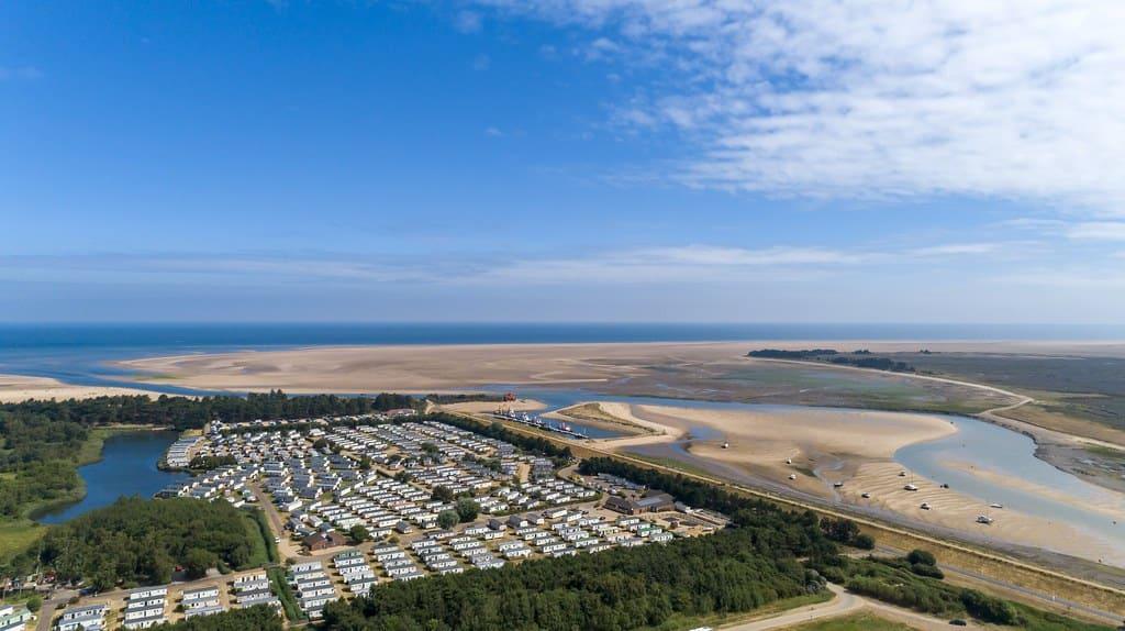 Aerial view of Pinewoods Holiday park next to a sandy beach and lagoon, under a bright blue sky with scattered clouds, conveying a peaceful, serene atmosphere.