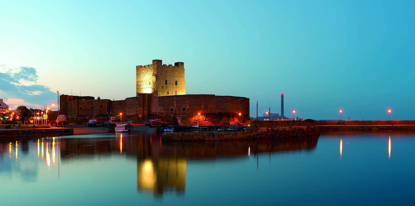 "Carrickfergus Castle at night lit up against a dusky sky with a reflection in the water in front"