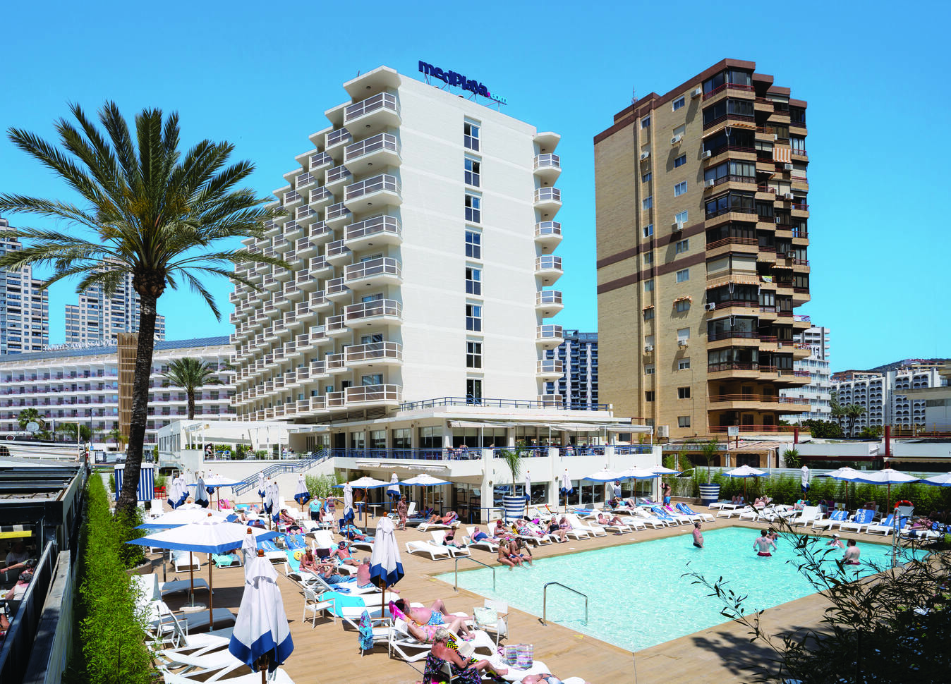 View of the pool area full of guests in and around the pool with the tall hotel buildings behind towering over on a clear sunny day.