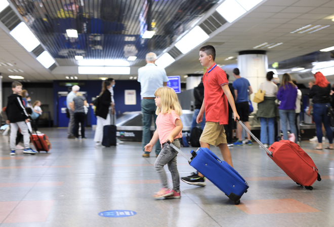 "Photograph shows two children in the foreground pulling their cases from the luggage carousels in arrivals, there are people in the background waiting for their luggage"
