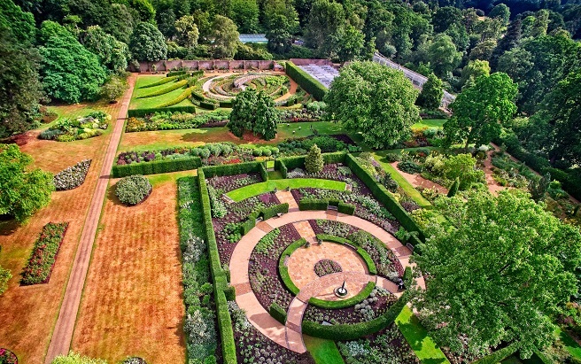 "Aerial view of The Savill Garden. There are many different layouts of plants and trees in different locations. At the top there is a spiral shape. There are trees surrounding the whole outside."