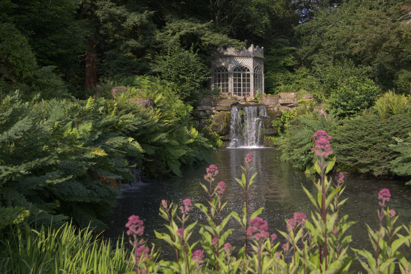 A view over the water and through the greenery of the Gothic Summer House with closed doors on the Long Walk.
