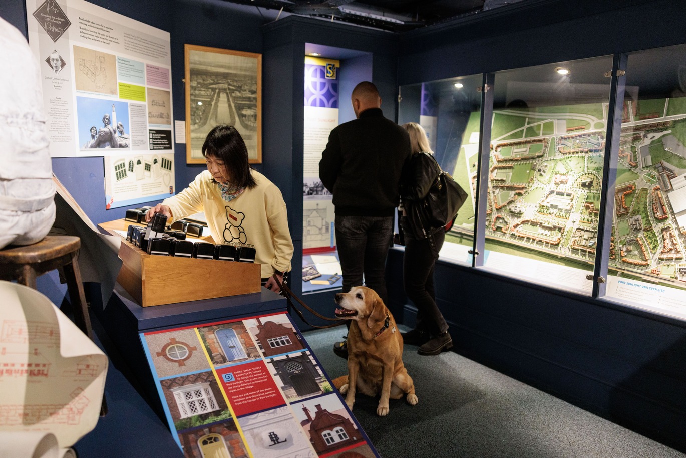 A woman uses a tactile exhibit while holding a harness for a guide dog. Other visitors view displays and a large architectural model in a museum setting.