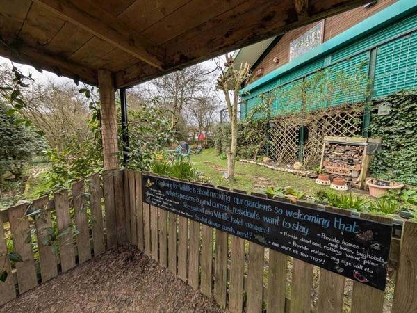A view from a wooden shelter shows a garden area with plants and trees beside a building, with a sign on the fence displaying a welcoming message about creating inclusive outdoor spaces