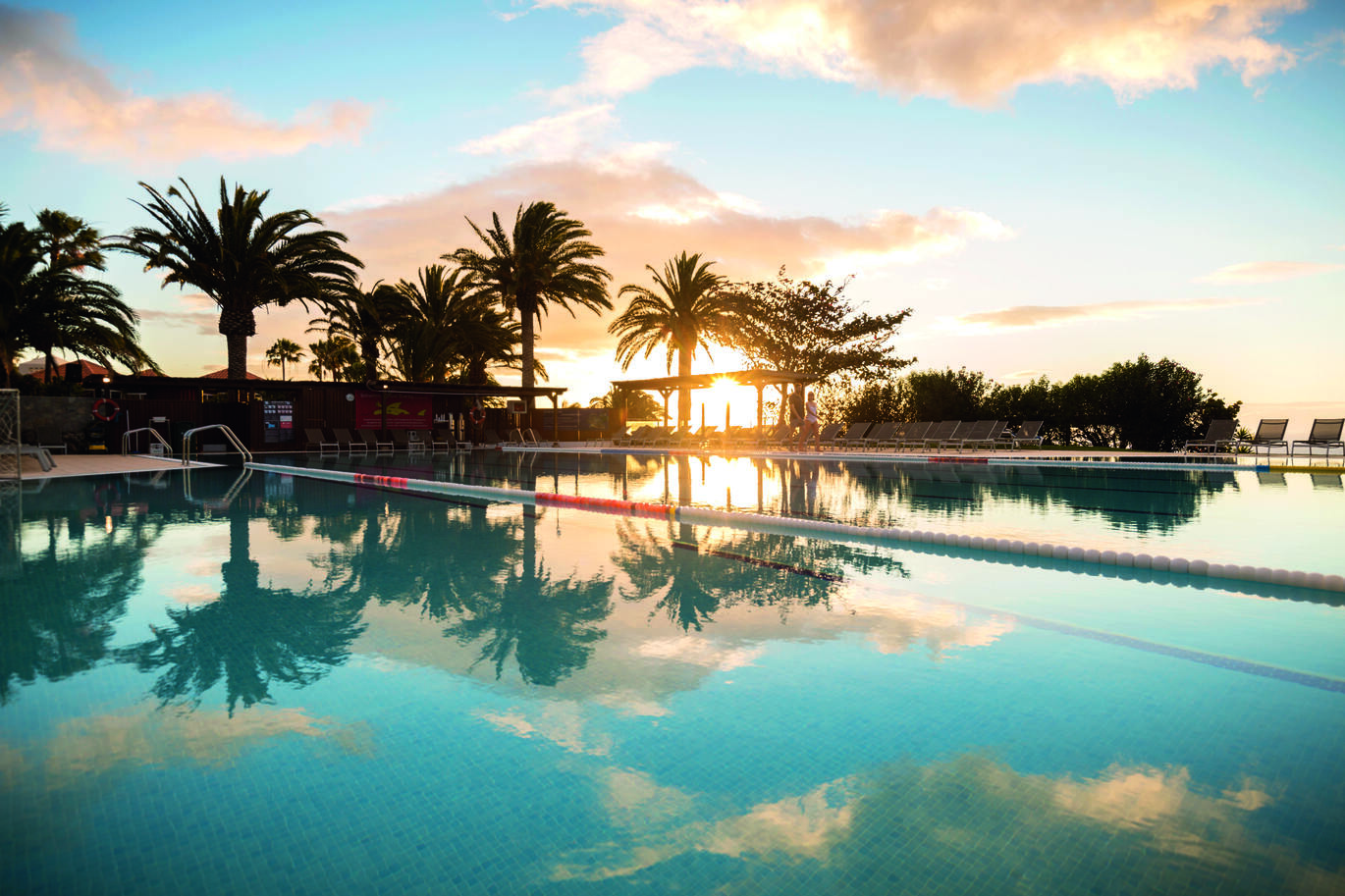 View from the pool side in the sunset surrounded with sun beds and trees reflecting in the pool with the slightly cloudy sky above.