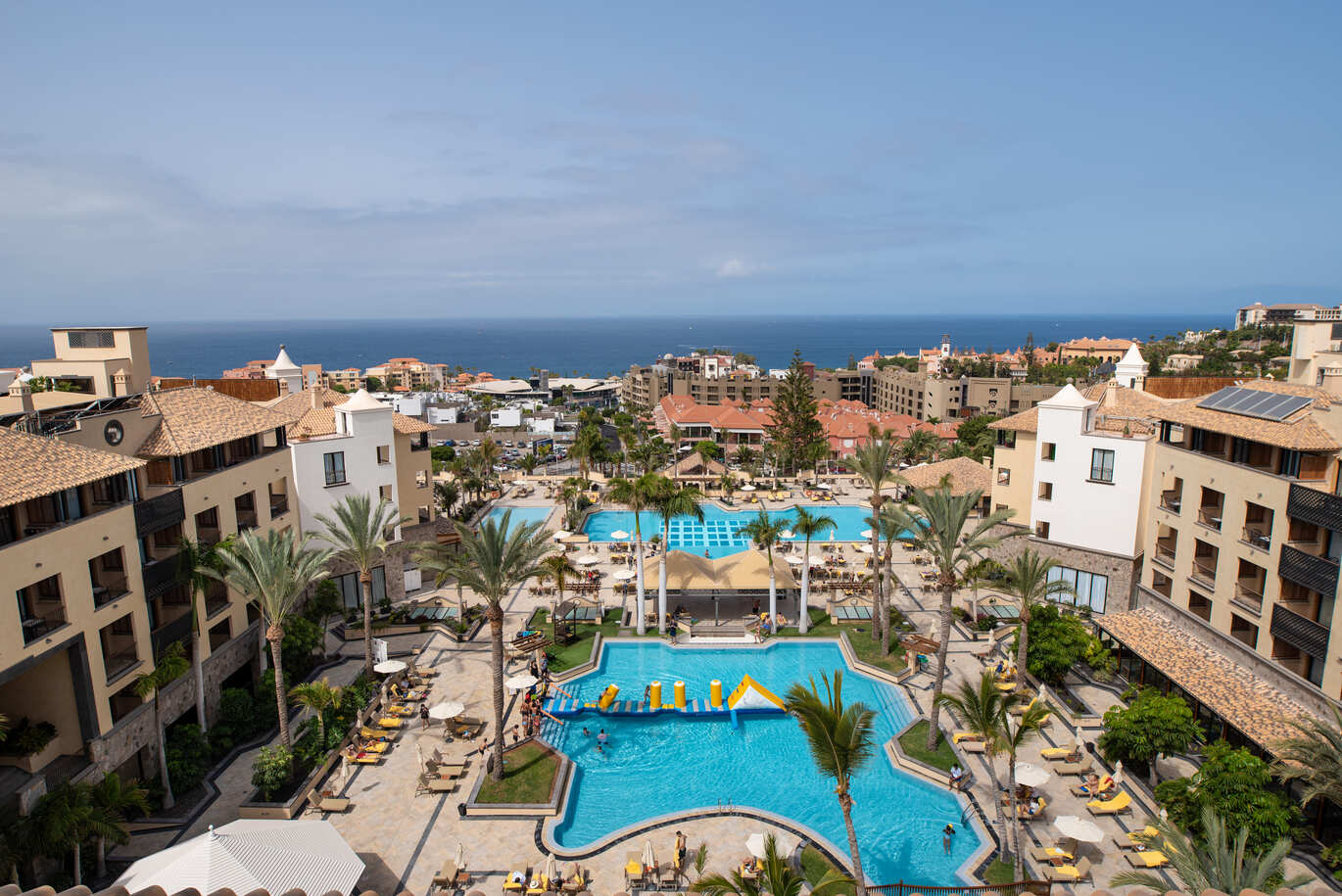 Aerial view of the hotel complex with the pool in the middle with an inflatable obstacle course on the water and sun loungers surrounding. With other buildings behind and the beach and sea beyond..