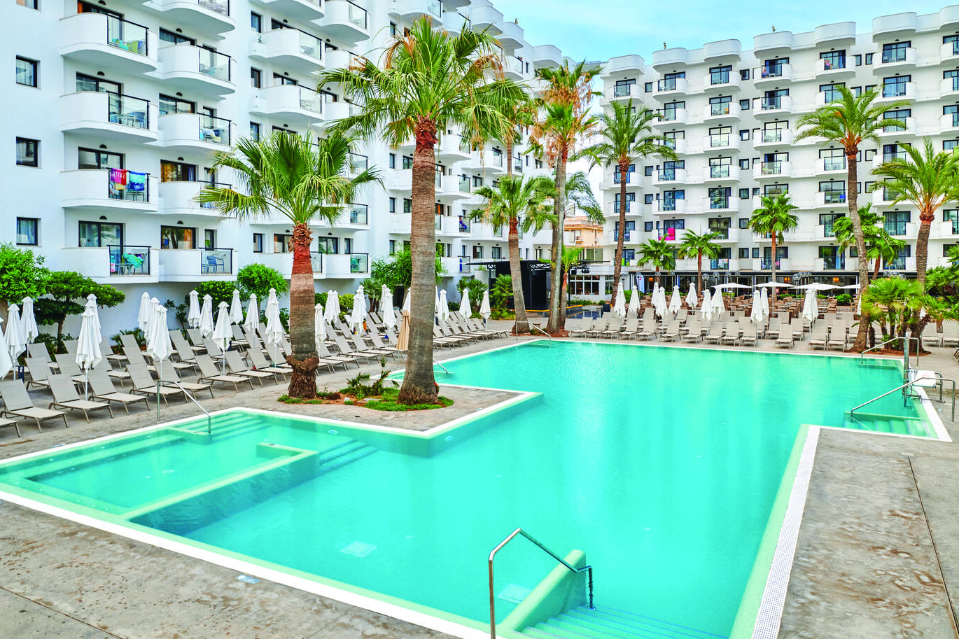 View of the pool with many sun loungers and umbrellas surrounding the pool with palm trees dotted around. Behind is the large hotel building with clusters of hotel room balconies on an overcast day.