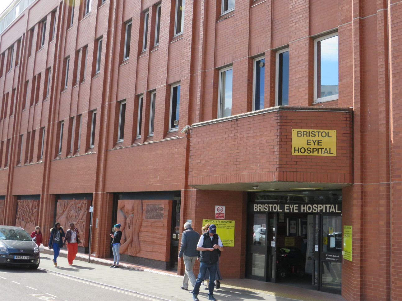 Bristol Eye Hospital yellow sign on the front of a large brick windowed building above the entrance with people walking past on the pavement and a car parked out the front.