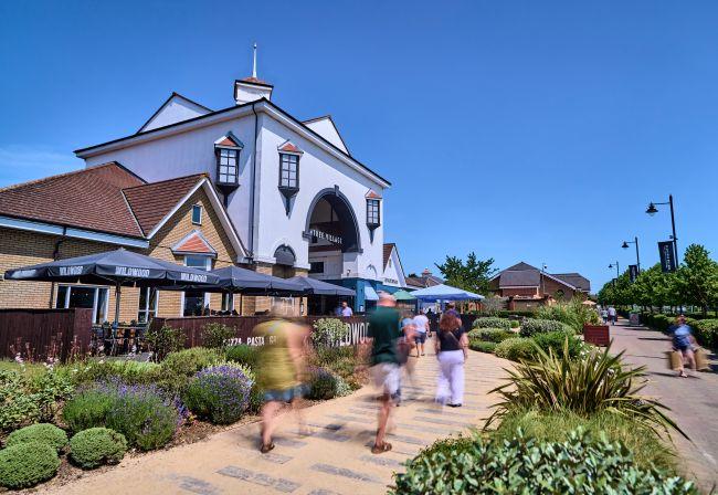 "Shoppers walk past landscaped paths and outdoor seating near the main Braintree Village entrance on a sunny day."