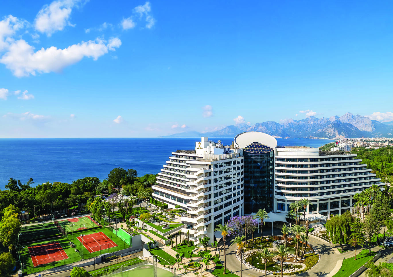 Aerial view of the hotel complex with tennis courts next to the large, white hotel building, surrounded by many trees with the sea behind and large mountains in the background.