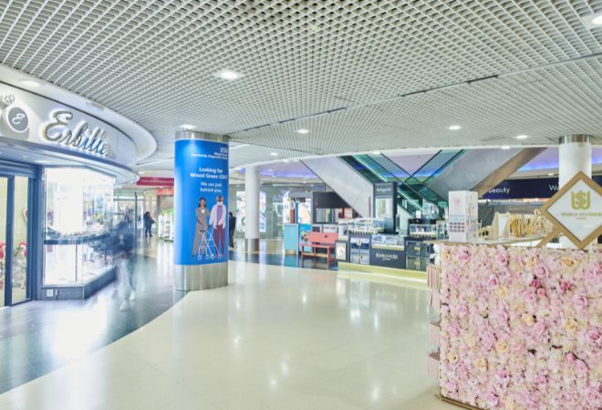 An indoor shopping mall with various retail stores. On the left, there is a store with the sign “Exhibit,” featuring a display window and a recessed entrance. The center of the image shows an open area with promotional stands, including one with a blue banner that has text and images of two individuals standing next to each other. To the right, there is a kiosk adorned with pink flowers, possibly for aesthetic or promotional purposes.