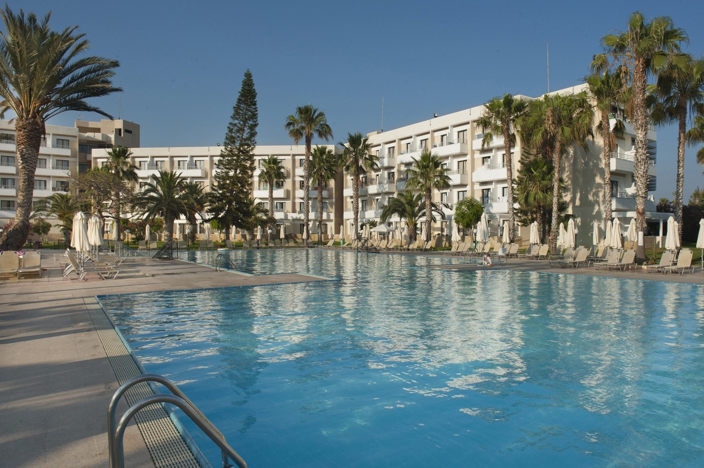 A view from the back corner of the blue pool surrounded by sun loungers and umbrellas with trees surrounding and the hotel building behind on a lovely clear blue day.