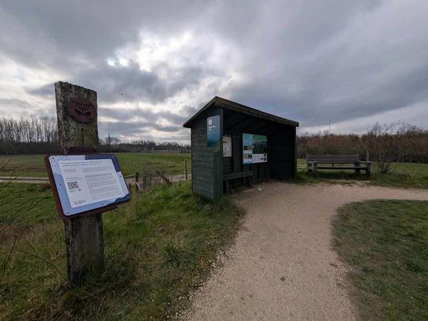 A scenic view of a nature trail with a wooden shelter and informational signs under a cloudy sky