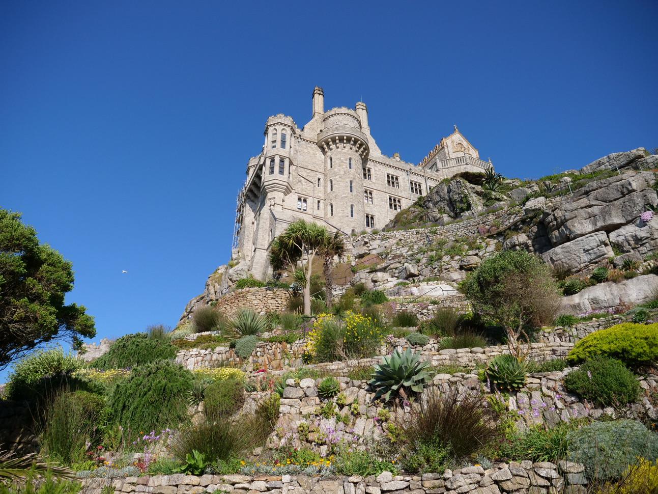 View of the terraced gardens and medieval castle on top of a steep hill made up of rocks with trees and greenery growing from them.