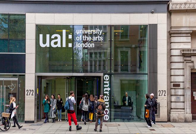 “Glass-fronted entrance of University of the Arts London with large white text ‘ual: university of the arts london’ on the upper section and ‘entrance’ vertically on the right. Several people are standing and walking on the pavement outside.”
