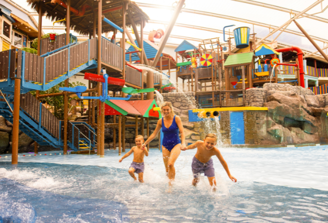 "Adult and children running through shallow water in a colourful indoor waterpark play area"