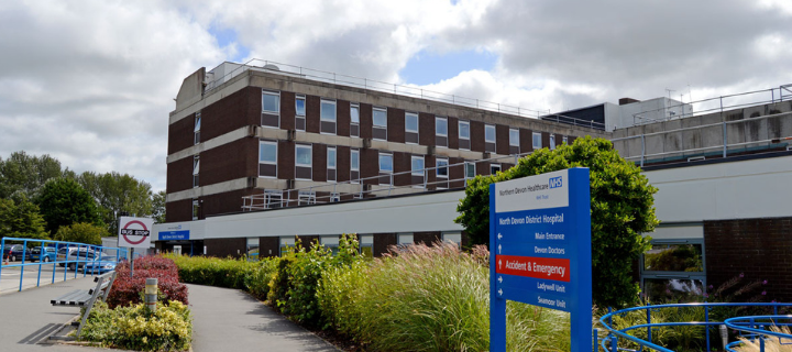 Blue signage showing arrowed directions around the hospital and a brown-brick multistorey hospital int eh background.  Pavement walkways shown and a car park on the left.